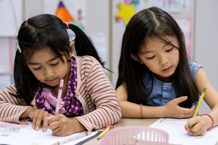 Two young students write on papers together at a desk in a classroom.